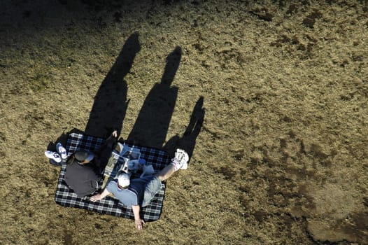 couple is sitting on rug having picnic, big shadows, sun must be very low, grass is dry