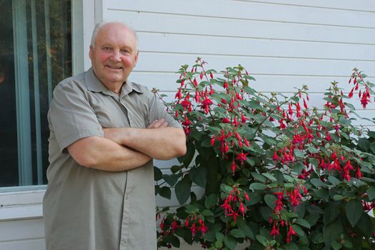 Old man - grower of flowers next to flower bush in summer day.