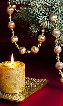 On a red background, a burning candle, fir branch and a ball chain.
