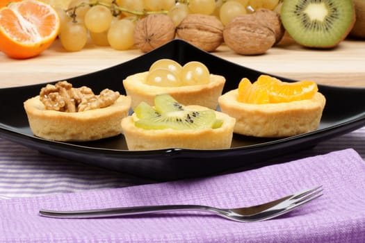Close-up of some mini fruit tarts served on a black plate with dessert fork. Selective focus.