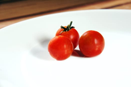 Three bright red ripe cherry tomatoes on a crisp white porcelain dinner plate placed on a wooden table.