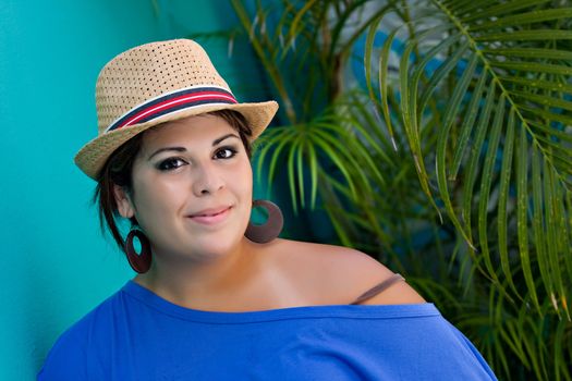 An attractive young Hispanic woman smiling with her hat and outdoors by some tropical foliage.