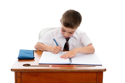 A young boy wearing a school uniform sitting at desk writing in a book.  White background.