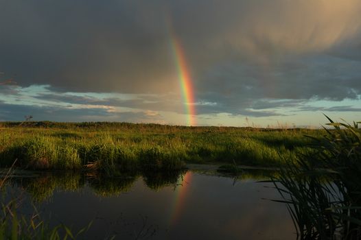 Evening rainbow above a small reservoir among a deserted field.
