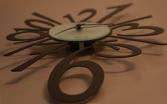Large abstract wall clock with modern black numbers and hands. Clock is resting against a tan background.