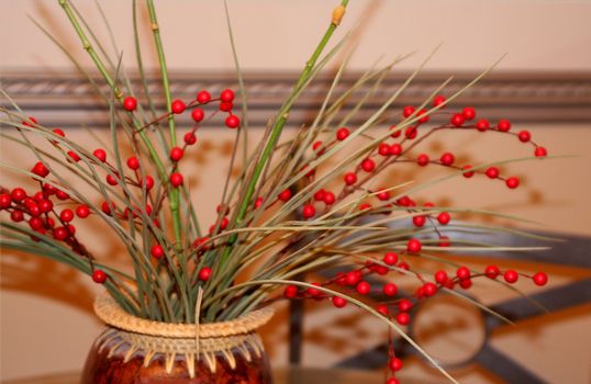 Vase of holly berries and greenery on kitchen table with chair in background. Wrought iron chair and chair rail behind image.