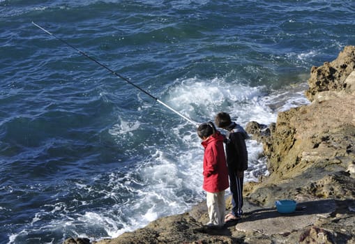 Two young fishermen on rocks with a blue sea