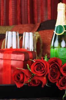 Beautiful long stem red roses with gift, wine and glasses in a breakfast tray on a bed.