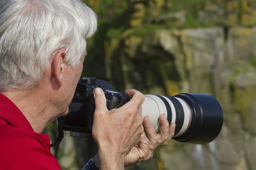 Mature man in red shirt with long lens mounted on camera looking out over natural, rocky area