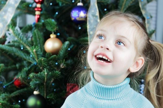 Excited little girl sits expectantly in front of a decorated Christmas tree.