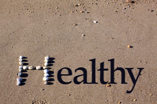 An upper cased letter 'H' formed by beach pebbles on the sand. The word healthy is spelt out by the addition of shadowed text.