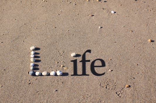 An upper cased letter 'L' formed by beach pebbles on the sand. The word Life is spelt out by the addition of shadowed text.