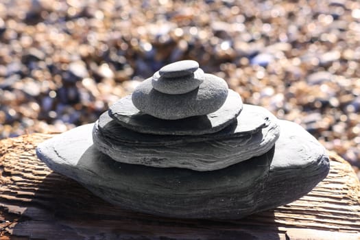 Pieces of gray slate stacked with gray pebbles piled on top, set on a block of drift wood. Slate has been shaped by the washing sea. A shingle beach set in soft focus to the background.