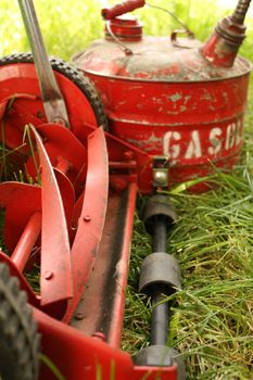 An environmentally friendly red push mower beside an old-fashioned red can of gasoline, both sitting in a lawn that needs cutting or mowing.  