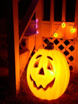 Halloween decorations: an illuminated bright orange pumpkin jack-o-lantern with a big toothy grin and some smaller pumpkin lights and purple lights behind it. Steps up to a white-painted deck are illuminated in the glow.