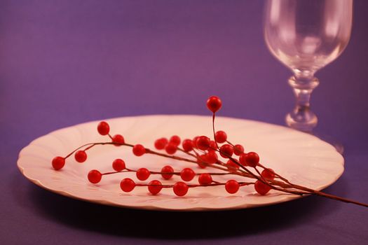 A white dinner plate with red holly berries on a blue background. A blurred piece of stemware is in the background. Holiday dinner plate for the season.