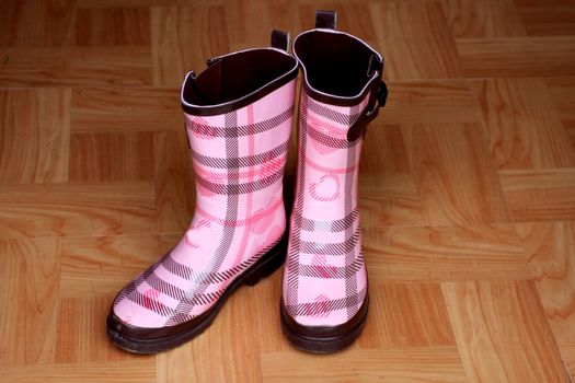 Adorable pink rain boots on wooden floor next to door. Women’s footwear for the rainy and cold season. Outdoor boots for rain.
