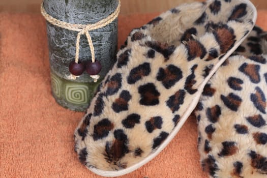 Pair of animal print leopard slippers for spa. Candle in background next to slippers. Items on orange towel for relaxation and spa treatment.