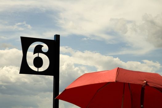 black number six, red umbrella in foreground, cloudy sky ind background