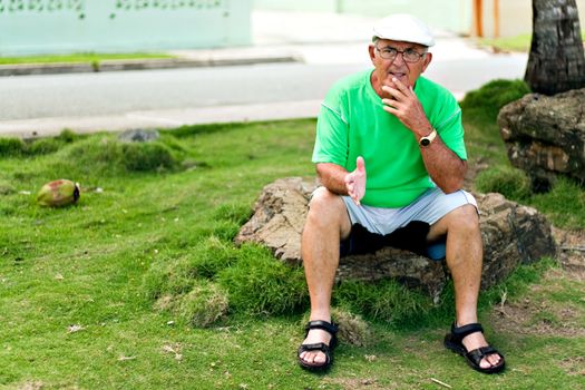 A older Hispanic senior citizen man sits outdoors in a tropical setting. 