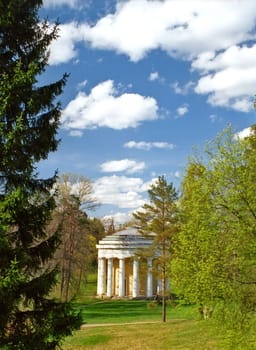 A beautiful classical white rotunda with the columnes is circled of trees in the spring park