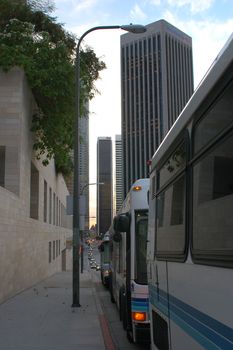 View of downtown LA along the sides of some local buses along a sidewalk with some skyscapers in the distance.