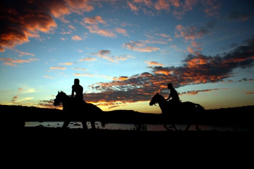 Silhouette of two horseriders on the background of a cloudy sky at sunset.
