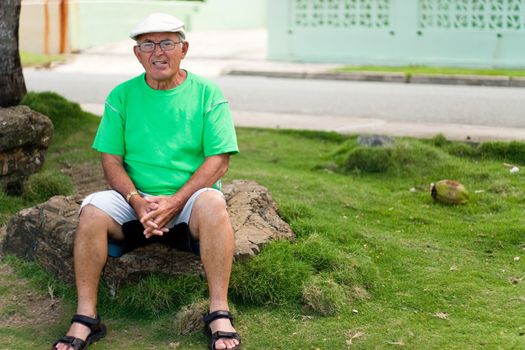 A older Hispanic senior citizen man sits outdoors in a tropical setting. 