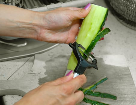 female hands peeling green fresh cucumber with peeler in kitchen