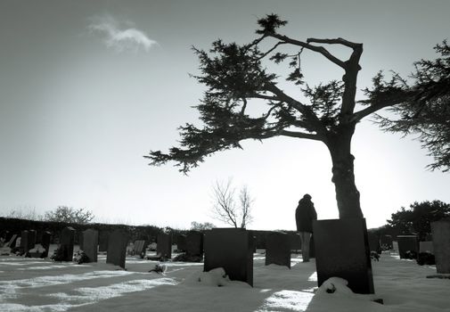 Lonely figure standing amongst tombstones in snowy cemetery.