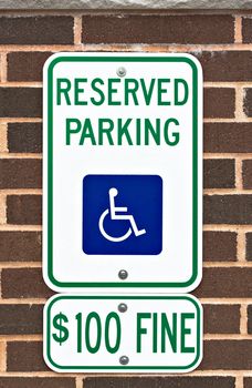 A reserved parking sign on a brick wall. There is a picture or symbol of a wheelchair on the sign along with a message for a one hundred dollar fine.
