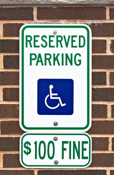 A reserved parking sign on a brick wall. There is a picture or symbol of a wheelchair on the sign along with a message for a one hundred dollar fine.