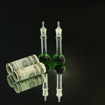 Two laboratory beakers with green liquid stand next to American currency on a mirror base with light flare on green beakers to enhance suggestion of successful investments in science;