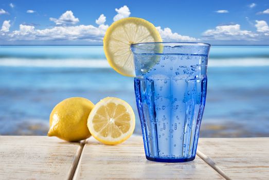a Blue glass with sparkling water and lemon on a wooden deck overlooking a tropical beach