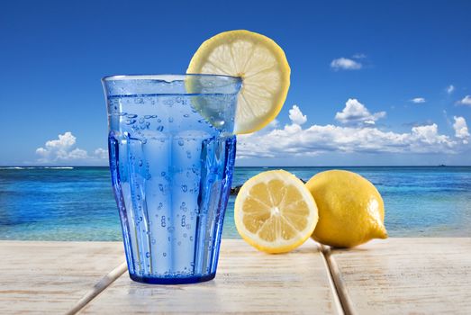 a Blue glass with sparkling water and lemon on a wooden deck overlooking a tropical beach