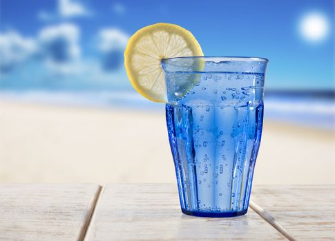 a Blue glass with sparkling water and lemon on a wooden deck overlooking a tropical beach - focus on the lemon