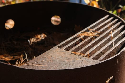 A rusting iron fire ring, filled with dried pine needles and fallen autumn leaves. A simple design adds extra functionality with a grilled shelf for some old fashioned barbequing.