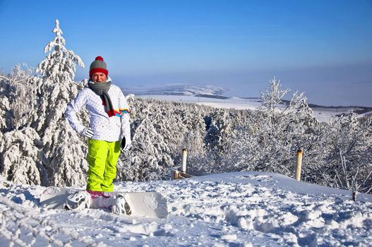 Beautiful woman with snowboard stands on top of the mountain. Against the backdrop of snow-covered pine trees and blue sky.