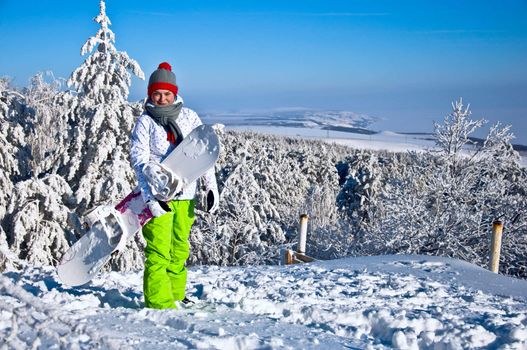 Beautiful woman with snowboard stands on top of the mountain. Against the backdrop of snow-covered pine trees and blue sky.