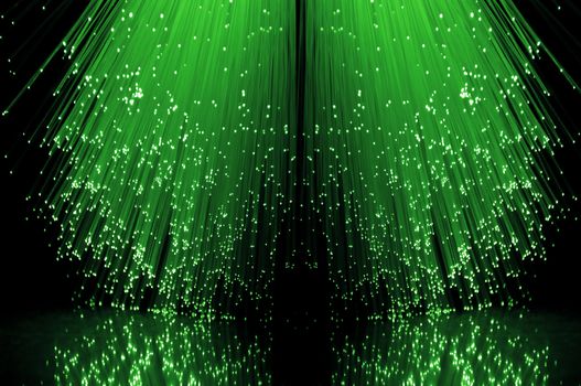 Low level angle capturing the ends of many illuminated green fibre optic light strands against a black background in reflecting into the foreground.
