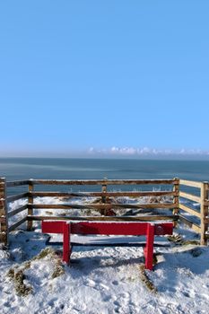 a seasonal snow frosty covered red bench with a sea view and blue cloudy sky background with copy space