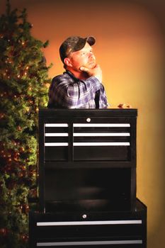 Adult male with thoughtful look leaning on black tool box with christmas tree in background