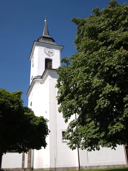 White Catholic Church in small town against blue sky with green trees around