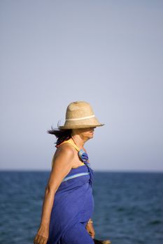 View of a middle-age woman walking on the beach with a hat.