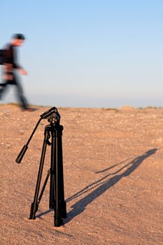 View of a tripod on the sand and a blured person walking by on the back.