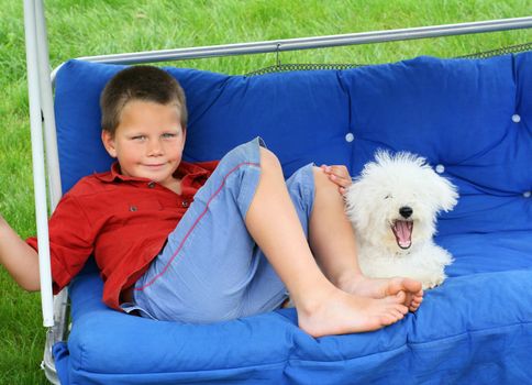 A blond smiling boy and a yawning puppy relaxing on a porch swing