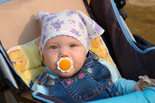 Pretty little girl with baby's dummy sit in carriage. In kerchief and jeans catsuit.