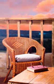 Wicker chair with pillow next to a wooden table with a book and reading glasses on it, with a view of a river at sunset in the background