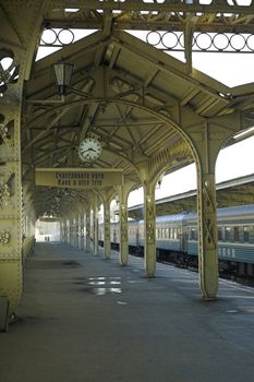 Railroad station platform with a hanging clock, "Have a nice trip" signboard, and two remote human profiles.