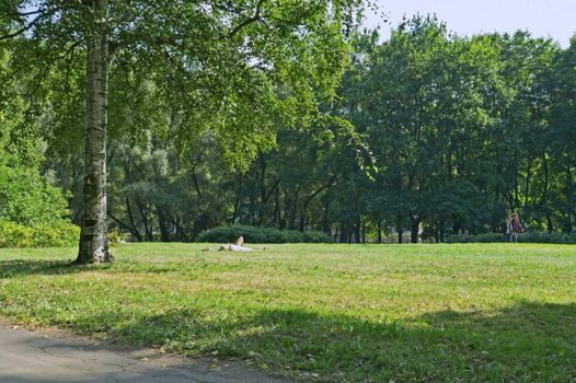 City park in summer and girl lying far off on grass and taking sunbathing.